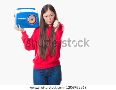 Young Chinese woman over isolated background holding vintage radio with angry face, negative sign showing dislike with thumbs down, rejection concept