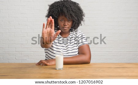 Young african american woman sitting on the table drinking a glass of milk with open hand doing stop sign with serious and confident expression, defense gesture