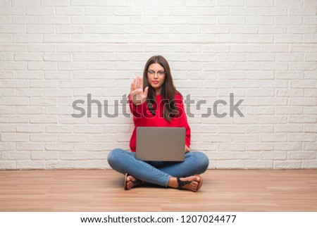 Young brunette woman sitting on the floor over white brick wall using laptop with open hand doing stop sign with serious and confident expression, defense gesture