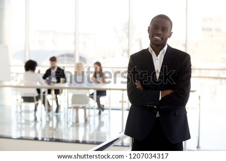 Portrait of smiling African American employee look at camera making picture in company hallway, confident black businessman or worker in suit standing posing for photo in office building