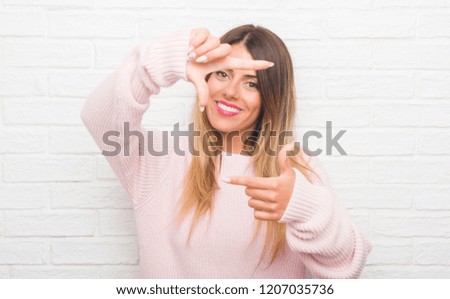 Young adult woman over white brick wall wearing winter outfit at home smiling making frame with hands and fingers with happy face. Creativity and photography concept.