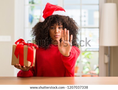 African american woman holding present wearing christmas red hat with open hand doing stop sign with serious and confident expression, defense gesture