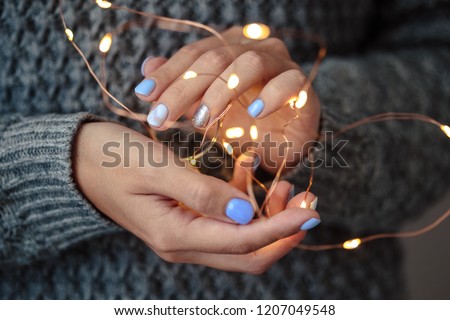 Gorgeous manicure, pastel tender color nail polish, closeup photo. Female hands hold a christmas light garland over grey knit background