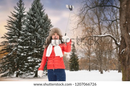 people, technology and leisure concept - happy woman in fur hat taking picture by smartphone selfie stick over winter forest background