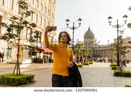 The young man waiting someone and does selfie, he dressed in a yellow sweater, a black raincoat or jacket, jeans, street and Kazanskiy cathedral on background, sunny day