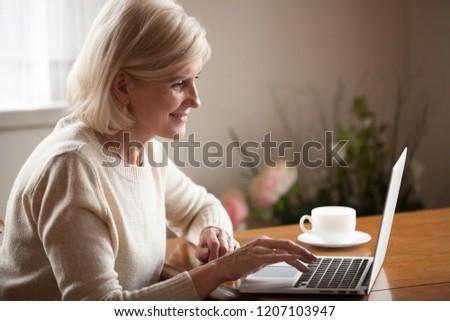 Excited aged woman using laptop, browsing internet at home, smart senior lady working at computer drinking coffee indoors, busy elderly female surfing web, checking mail or reading news online