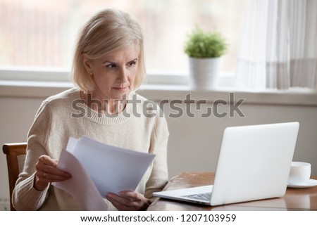 Serious aged woman holding documents, checking information at laptop online, concerned senior female managing bank insurance or loan papers, busy working at computer. Elderly and technology concept