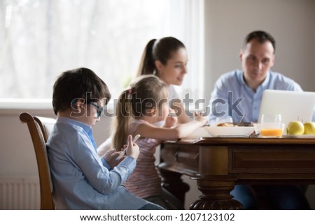 Family with little children sitting in kitchen during breakfast. Wife husband looks at laptop, focus on small son in glasses use cellular. Mobile phone overuse and addiction gadgets dependency concept