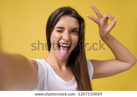 Pleasant attractive girl making selfie in studio and laughing. Good-looking young woman with brown hair taking picture of herself on bright yellow background.