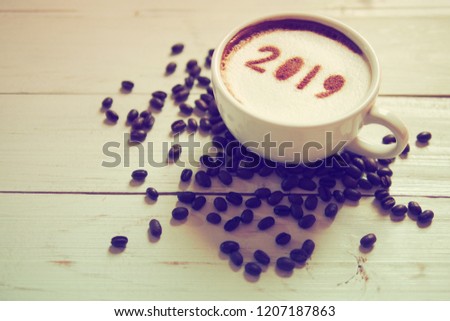 White cup of coffee with the number 2019 on frothy surface on white painted wooden background with coffee beans. Food art creative concept for active days in New Year 2019. (space for text)