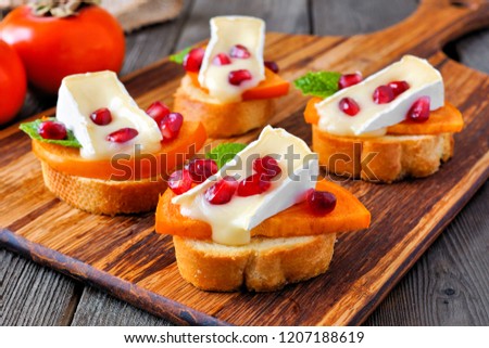 Holiday crostini appetizers with persimmons, pomegranates and brie cheese. Close up, side view scene on a wood background.