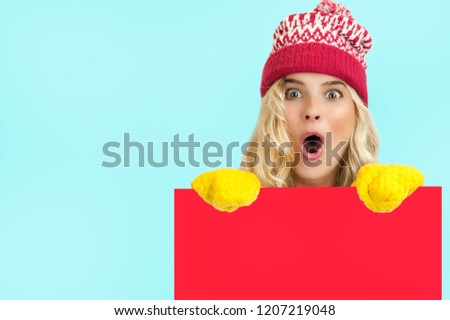 
A portrait of a very beautiful bright girl in Christmas clothes posing behind a red panel isolated on a blue background Holding a blank Christmas billboard