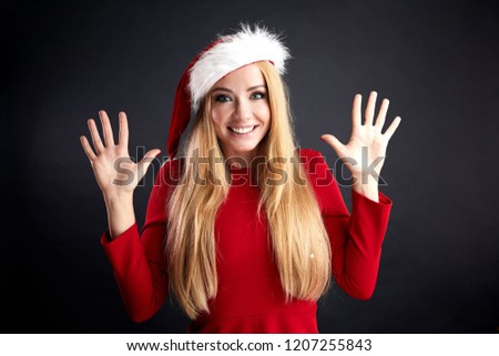 Excited young blonde girl with long hair wearing Santa hat and red elegant dress gesturing with two hands isolated over black background