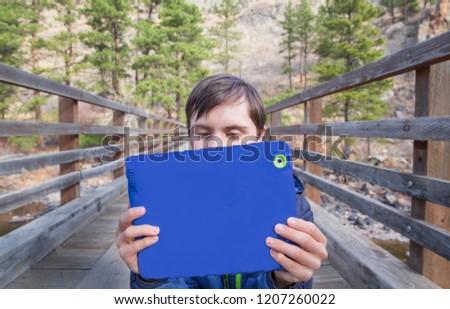 horizontal orientation color image of an attractive teenage boy with autism and down's syndrome on a bridge outside, using his tablet device