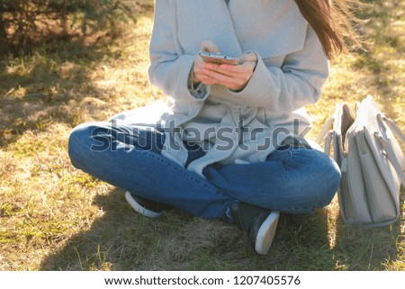 Young beautiful brunette woman sits on grass in spring park and types a message in mobile phone. Close-up hands.