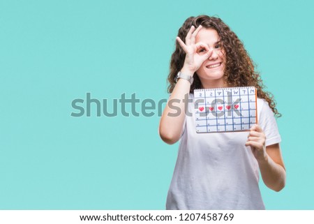 Young brunette girl holding menstruation calendar over isolated background with happy face smiling doing ok sign with hand on eye looking through fingers