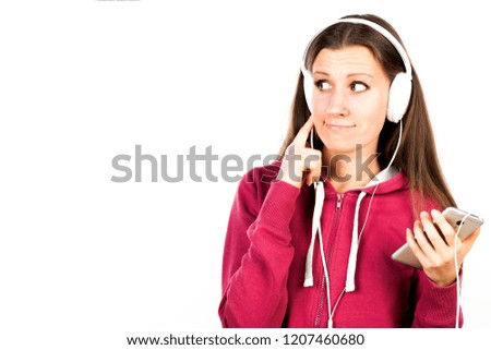 Young attractive smiling girl with long hair and pierced tongue listening the music by smartphone with headphones and making faces. Advertising concept isolated on abstract blurred white background