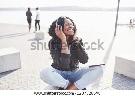 Portrait of a beautiful young pretty African American girl sitting on the beach or lake and listening to music in her headphones.