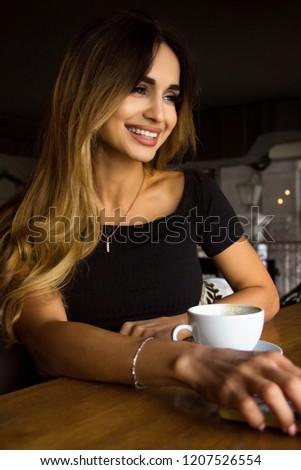 young happy pretty woman in cafe. female is smiling with white cup near her hand and looking away. girl is sitting   near window  on cafe hall background. lifestyle concept portrait