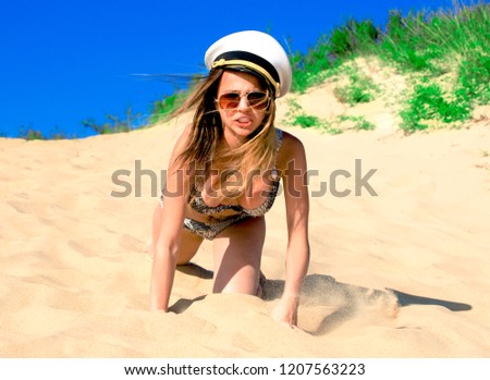 The girl in the captain's cap creeps along the sandy beach at her knees. Cute model in a black white swimsuit resting on a white beach.