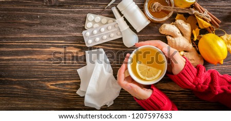 Medical care concept - hands holding cup with ginger honey and lemon tea with drugs, pills and spray on wood background Royalty-Free Stock Photo #1207597633