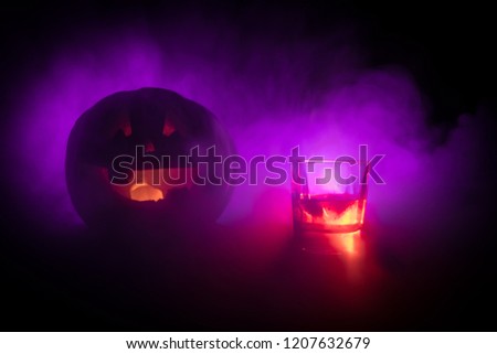 Halloween pumpkin with carved face and glass of whiskey with ice on a dark toned foggy background. Decorated. Selective focus