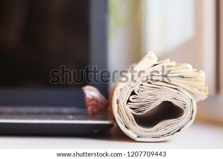 Rolled newspaper next to computer. Daily papers with news and open laptop. Tabloid journal and black screen of electronic device. Concept for researching and using different sources of information    