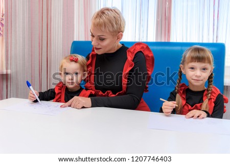 Photo portrait of mother and two cute girls draw on the table in the room. They are sitting right in front of the camera smiling and looking happy