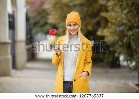 Fashion portrait pretty sweet young woman in yellow coat having fun with red lollipop heart over autumn street background