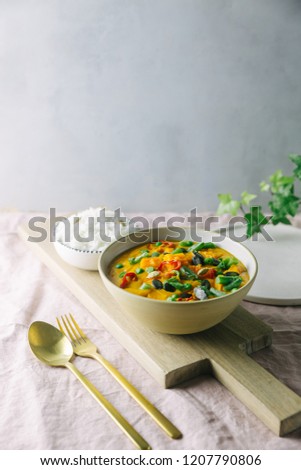 Vegan healthy red curry with pumpkin, green peas, green beans and chili served with jasmin rice on a chopping board. Pink, grey, subtle background.