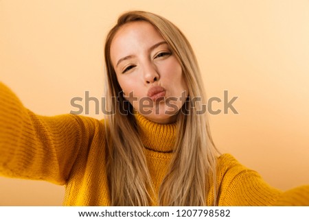 Portrait of a lovely young woman dressed in sweater standing isolated over yellow background, taking a selfie
