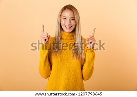 Portrait of a happy young woman dressed in sweater standing isolated over yellow background, pointing up at copy space