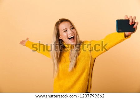 Portrait of a happy young woman dressed in sweater standing isolated over yellow background, taking a selfie