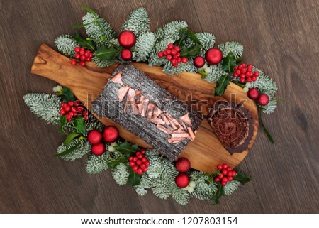 Christmas chocolate log yule cake with traditional winter flora of snow covered fir, holly and mistletoe on rustic wooden board and oak table background. 