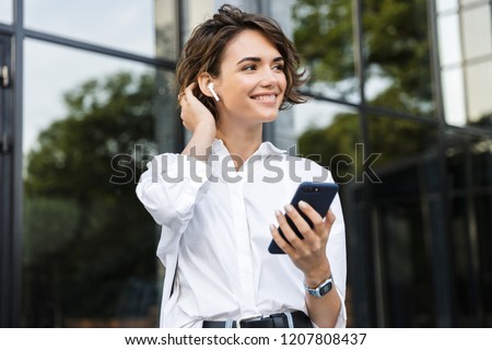 Smiling young woman in earphones standing outdoors at the street, talking on mobile phone Royalty-Free Stock Photo #1207808437