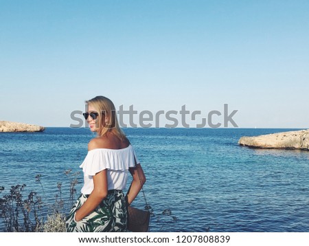 Back view of fashionable young female tourist standing on the rock, admiring beautiful and calm nature around. Pretty blonde woman smiles, posing at blue ocean, waiting for friend to make picture.