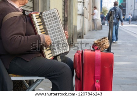Close Up of Old Beggar Woman Playng a Dirty Accordion in the Street near a Red Suitcase on Blur Background