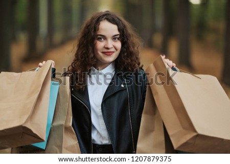 Photo of pretty happy woman with shopping bags in park, smiling curly girl enjoying of new purchase over autumn foliage background, fashion and style lifestyle, spending money concept