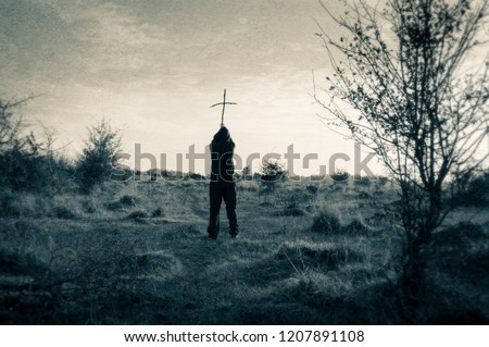 A lone hooded figure standing outside holding a wooden cross. silhouetted against the afternoon winter sun. With a vintage, grunge duo tone edit