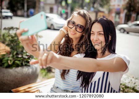 Two pretty thin girls with long dark hair,dressed in casual stye,sit at the bench and take a selfie,