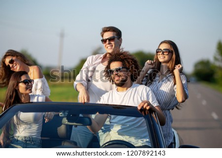 Stylish young girls and guys in sunglasses dressed in a white clothes are sitting  in a black cabriolet up on a sunny day.