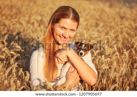 Young woman holding Jack Russell terrier puppy on her hands, both of them smiling, sunset lit wheat field in background.