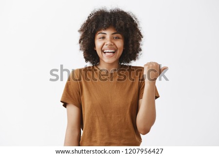 Waist-up shot of carefree joyful and attractive young dark-skinned female student in stylish t-shirt pointing right and smiling broadly while showing way or asking question about curious thing