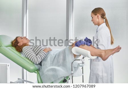 Side view portrait of young gynecologist in white lab coat and sterile gloves holding medical vaginal speculum. Red-haired woman with closed eyes lying in gynecological chair