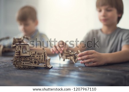 Laconic photo of two boys playing with wooden constructor and ready model standing on the table