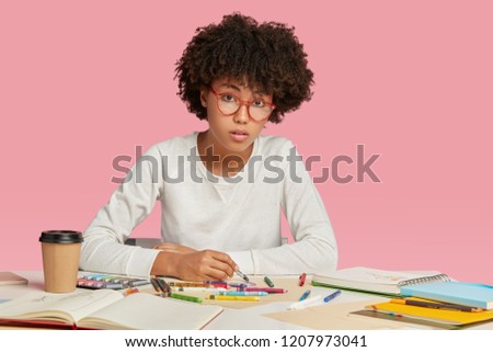 Female architect has Afro hairstyle, works on designing project, draws pictures in notebook looks seriosly at camera, wears spectacles, likes creativity, isolated over pink background, busy with work