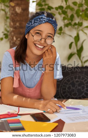 Indoor shot of pleased author with toothy smille, wears round big glasses for better vision, dressed in casual clothes, sits at workspace with documentation, writes creative ideas in notepad. Royalty-Free Stock Photo #1207973083