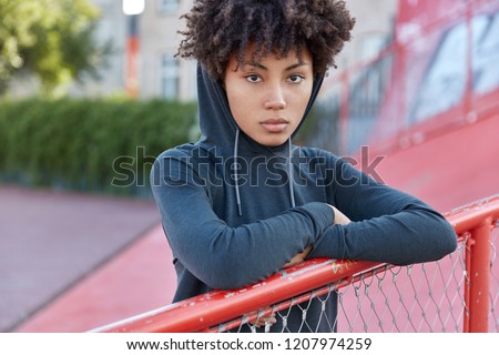 Cropped shot of serious dark skinned woman hipster has curly hair, dressed in sweatshirt with hoodie, leans at fence, spends free time on playground, enjoys leisure waits for friend to play basketball