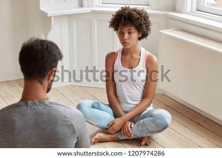 Indoor shot of mixed race boyfriend and girlfriend practice yoga, sit crossed legs, have serious talk about future, pose on wooden floor in spacious room. People, lifestyle and relaxation concept