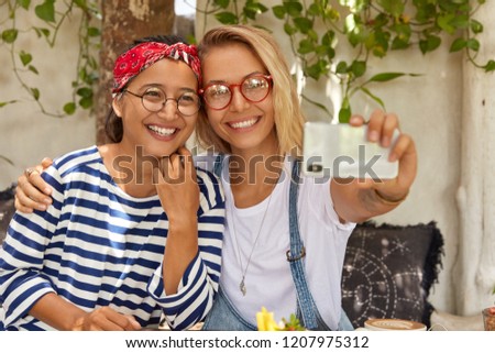 Positive friends of different nationalities make selfie portrait on modern smart phone, look at web camera, embrace each other, pose against cozy interior with green plant in background. Spare time